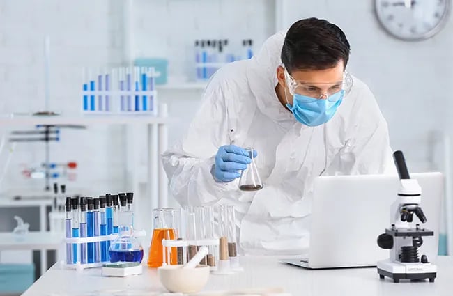 Lab tech holding beaker with fluid reviewing a laptop